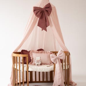 May include: A wooden oval crib featuring a light pink canopy with a large dark red gingham bow. Inside are a light pink ruffled pillow, a dark red gingham pillow, and a small white bear ornament. A matching pink bow is attached to the crib side.