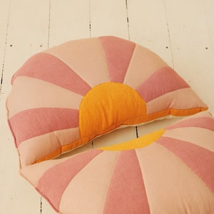 Könnte beinhalten: Zwei dekorative Kissen im Sonnenstrahl-Design. Die Kissen haben eine Halbkreisform mit abwechselnd rosa- und pfirsichfarbenen Strahlen, die von einer leuchtend orangefarbenen Mitte ausgehen. Die Kissen sind aus Stoff und liegen auf einer weißen Holzoberfläche.