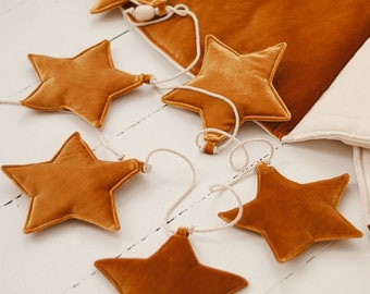 May include: A garland of six plush velvet stars in a mustard yellow colour, connected by a white string. The stars are arranged in a staggered pattern, with the ends of the string hanging down.