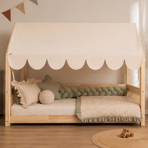 Könnte beinhalten: Ein helles Holzbettgestell in Hausform mit einem cremefarbenen Stoffdach mit gewelltem Rand. Das Bett ist mit einer cremefarbenen Matratze, einer gemusterten Steppdecke mit salbeigrünem Rüschenrand, einem geflochtenen Bettschutz und Kissen ausgestattet.
