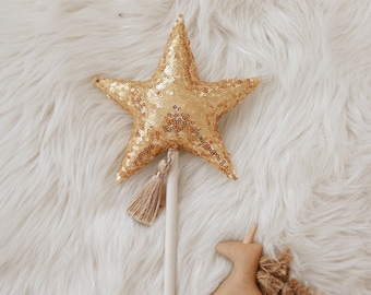 Könnte beinhalten: Ein goldener sternförmiger Zauberstab mit einem Holzstab und einem braunen Quasten. Eine Holz-Einhorn-Figur mit einem braunen Quasten-Schwanz liegt auf einer weißen flauschigen Oberfläche.
