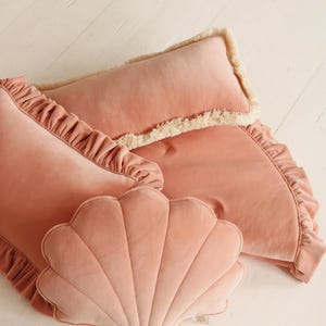 May include: Three pink velvet pillows, one shaped like a seashell, one with a ruffled edge, and one long pillow with a white fringe.