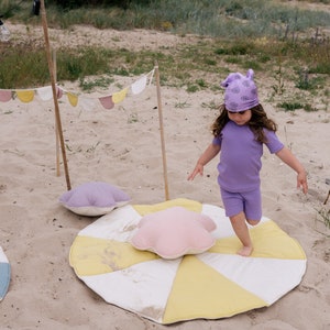 Puede incluir: Un niño con una camisa y un gorro morados juega en una alfombra de juego en forma de estrella amarilla y blanca en una playa de arena. Hay otras alfombras de juego de colores y una hilera de banderas en el fondo.