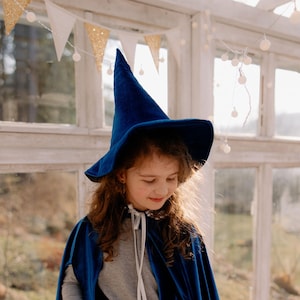 Puede incluir: Un disfraz de bruja azul de terciopelo para niños, que incluye un sombrero puntiagudo y una capa. El atuendo presenta una falda de tul en capas y una cintura con lentejuelas plateadas. El niño sostiene una varita en forma de estrella.