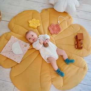 May include: A baby lies on a yellow flower-shaped play mat with a variety of textures. The mat has a pink flower, a yellow flower, and a white crescent moon attached. The baby is holding a wooden toy.