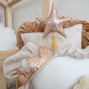 Peut inclure: Un oreiller blanc avec un bord à volants et une couronne en paillettes dorées sur le dessus. L'oreiller est posé sur un lit avec d'autres oreillers et une étoile dorée.