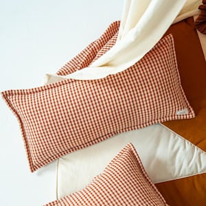 May include: Two rectangular pillows with a red and white gingham pattern. The pillows are placed on a white and brown patterned surface, with a cream-colored fabric draped over them. The pillows have a simple, classic design.