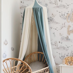 Puede incluir: Una escena de guardería con una cuna de ratán marrón claro, una almohada gris y un colchón blanco. Un dosel en crema y azul cuelga sobre la cuna. El papel pintado tiene un patrón de vida marina. También se ven una mesita de noche blanca y una silla pequeña.