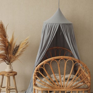 May include: A light gray canopy draped over a natural woven rattan bed frame with a white and gray patterned bedding. A gray velvet pillow sits on the floor in front of the bed.