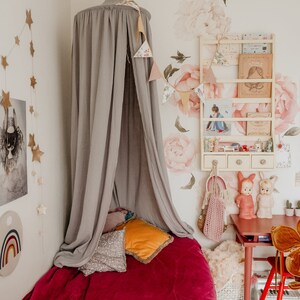 May include: A gray canopy bed with a red velvet blanket and pillows. The bed is in a room with a white wall and a floral wallpaper. There is a small table with a red chair and a white shelf with books and toys.