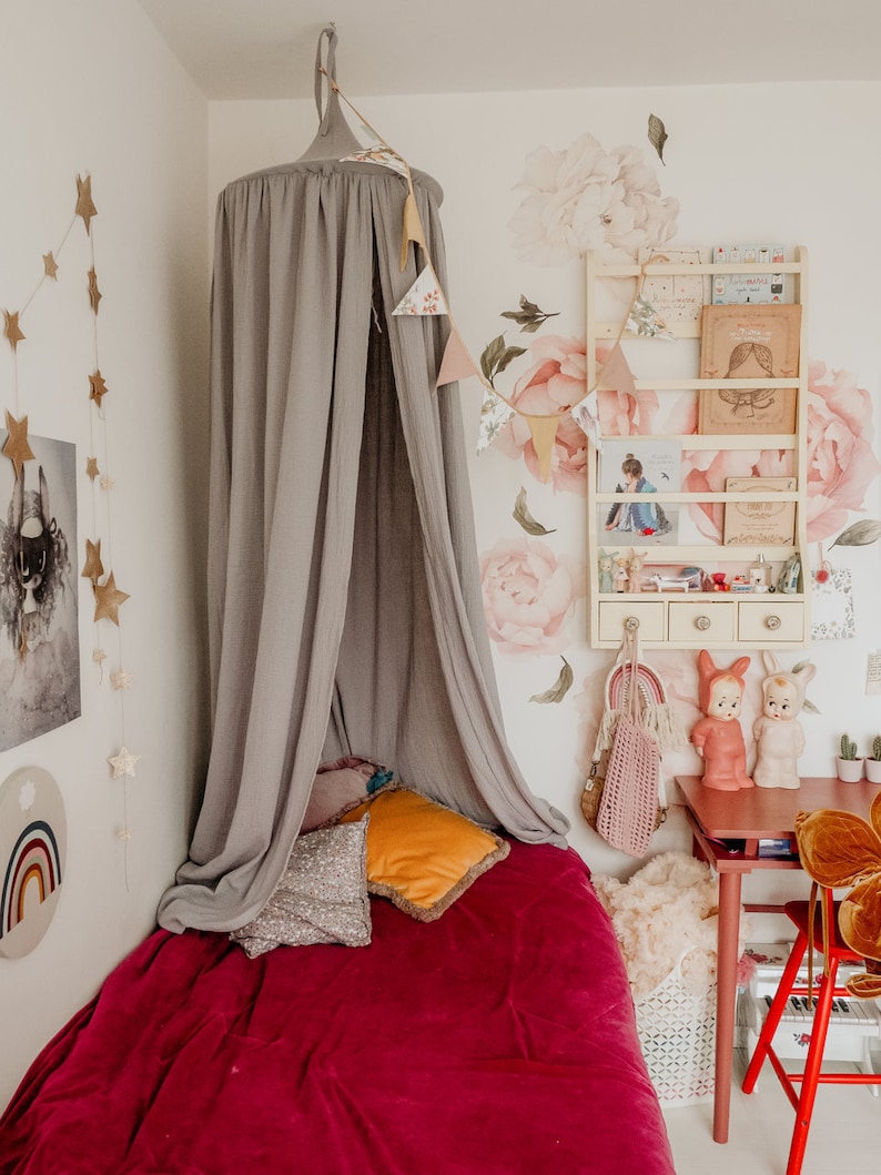 May include: A gray canopy bed with a red velvet blanket and pillows. The bed is in a room with a white wall and a floral wallpaper. There is a small table with a red chair and a white shelf with books and toys.