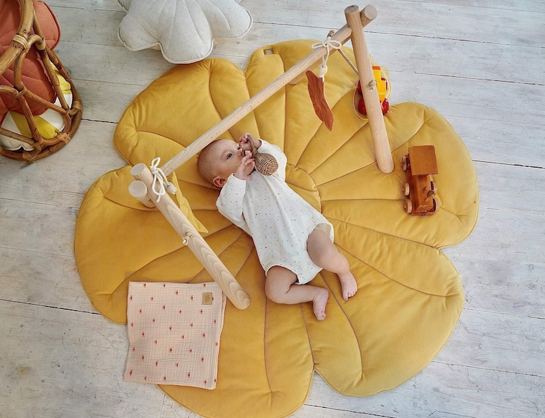 May include: A baby lies on a yellow flower-shaped play mat with a wooden play gym overhead. The play gym has a wooden toy car, a wooden toy train, and a wooden toy hanging from it.
