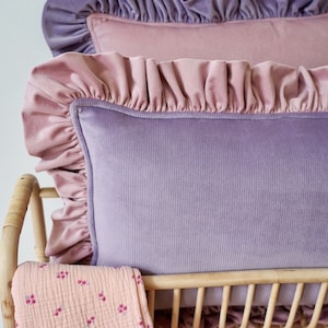 May include: Two ruffled velvet pillows in shades of pink and purple, resting on a woven wicker cot with a pink floral patterned blanket.