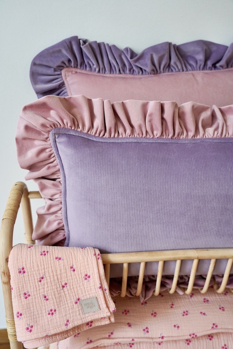 May include: Two ruffled velvet pillows in shades of pink and purple, resting on a woven wicker cot with a pink floral patterned blanket.