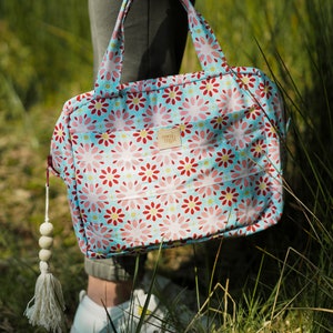 Könnte beinhalten: Eine Tragetasche mit Blumenmuster mit hellblauem Hintergrund und rosa und roten Blumenmuster. Die Tasche hat passende Griffe und ein kleines Etikett mit der Aufschrift "moi. miji". Ein weißer Quaste mit Holzperlen hängt am Reißverschluss.