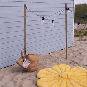 May include: A yellow flower-shaped play mat on a sandy beach with a brown woven tote bag and a string of lights hanging from wooden posts.