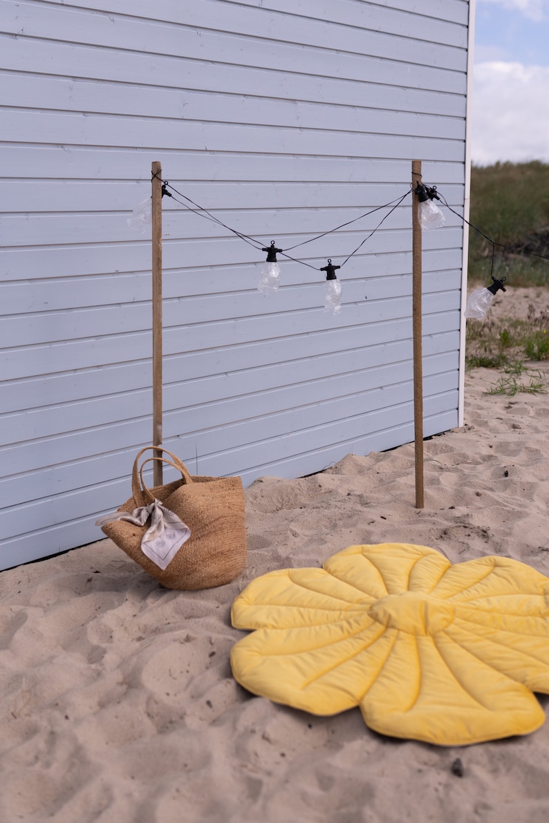 May include: A yellow flower-shaped play mat on a sandy beach with a brown woven tote bag and a string of lights hanging from wooden posts.