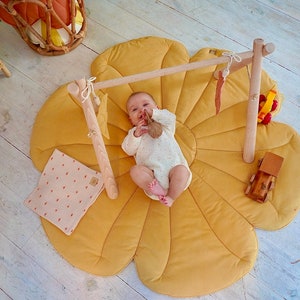May include: A baby lies on a yellow flower-shaped play mat with a wooden play gym overhead. The baby is holding a knitted toy.