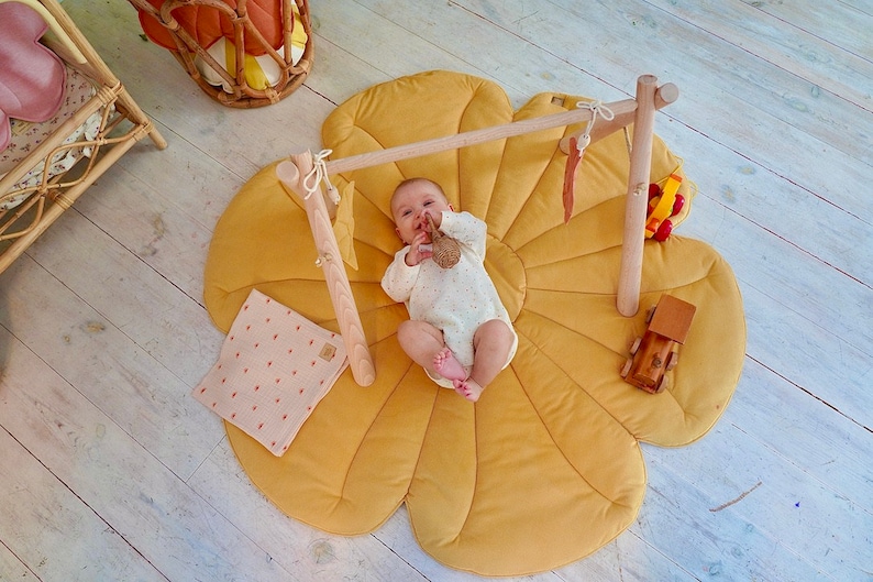May include: A baby lies on a yellow flower-shaped play mat with a wooden play gym overhead. The baby is holding a knitted toy.