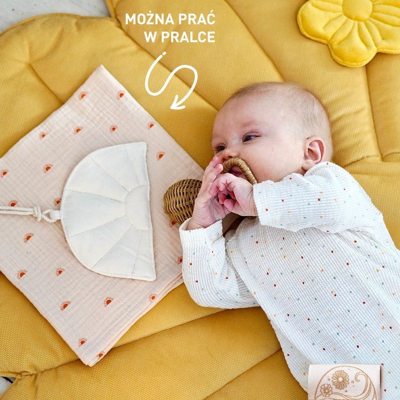 May include: A baby lying on a yellow and pink play mat with a white muslin swaddle blanket with a small white crescent moon shape sewn on it. The baby is wearing a white long-sleeved onesie with colourful polka dots and is holding a woven basket toy. The text "MOŻNA PRAĆ W PRALCE" is visible in the top centre of the image.