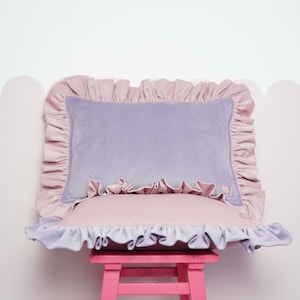 May include: A lavender velvet cushion with a pink frilled trim sits on a pink stool. The cushion has a pink frilled trim around the edges.