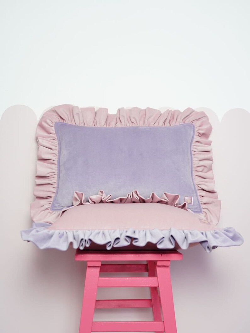 May include: A lavender velvet cushion with a pink frilled trim sits on a pink stool. The cushion has a pink frilled trim around the edges.