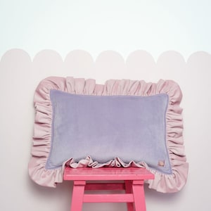 May include: A lavender velvet cushion with a pink frilled trim sits on a pink wooden stool against a white wall with a pink scalloped border.