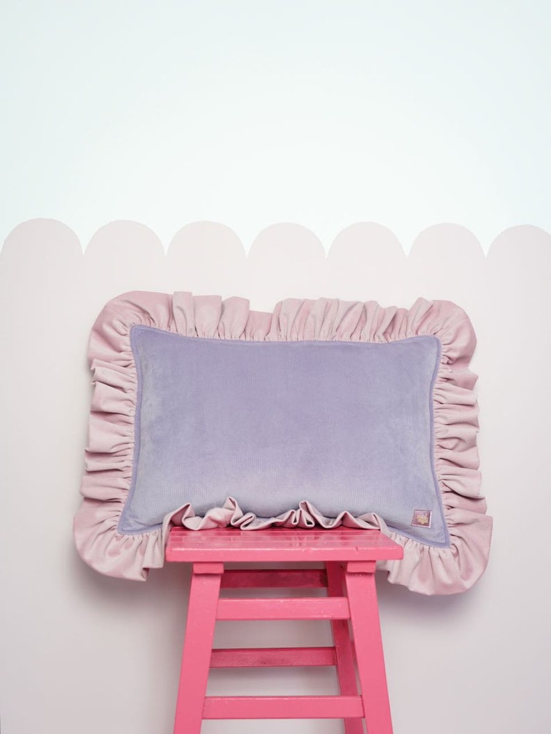 May include: A lavender velvet cushion with a pink frilled trim sits on a pink wooden stool against a white wall with a pink scalloped border.