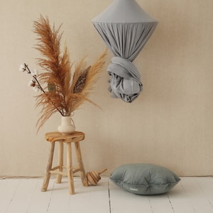 May include: A gray fabric canopy hanging from the ceiling, a wooden stool with a vase of dried pampas grass, and a gray quilted pillow on a white wood floor.