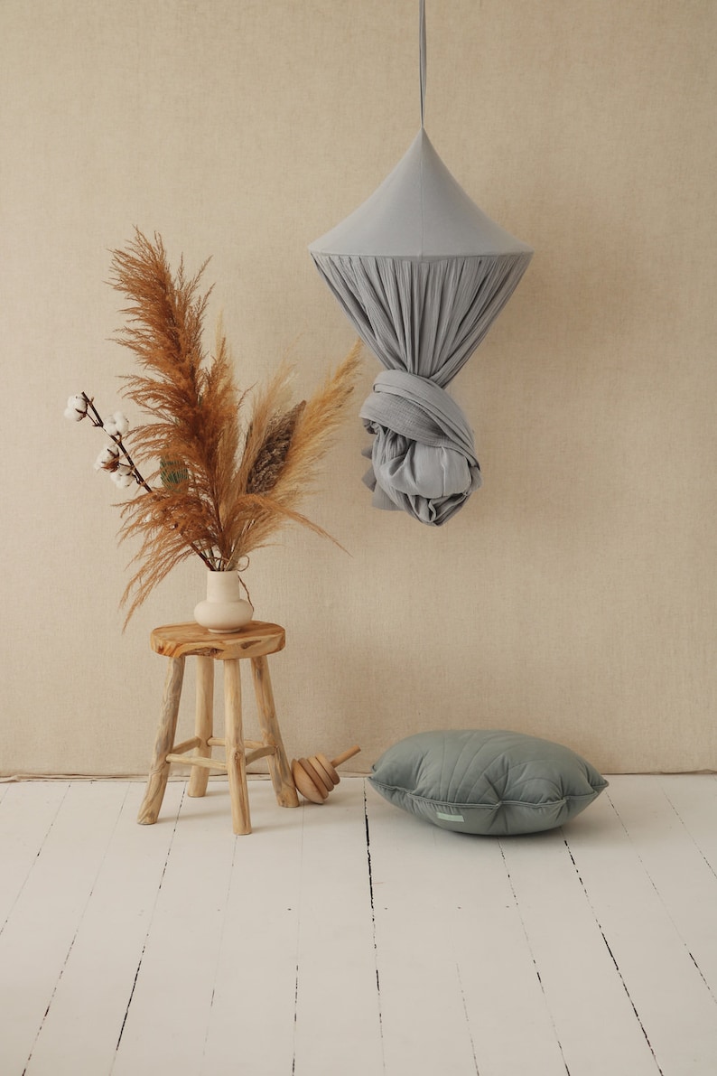 May include: A gray fabric canopy hanging from the ceiling, a wooden stool with a vase of dried pampas grass, and a gray quilted pillow on a white wood floor.