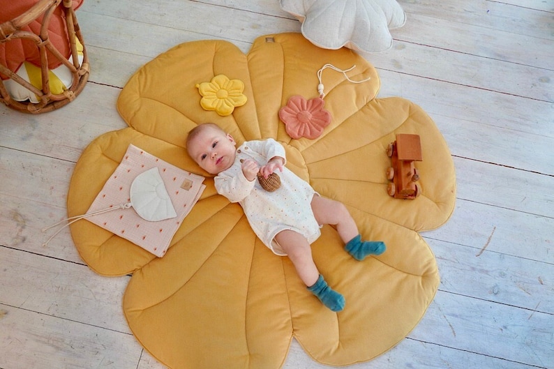 May include: A baby lies on a yellow flower-shaped play mat with a variety of textures. The mat has a pink flower, a yellow flower, and a white crescent moon attached. The baby is holding a wooden toy.