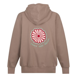 以下が含まれることがあります： 薄い茶色のフード付きスウェットシャツ。背面にピンクとグリーンの刺繍の花と「peace and bloom」の文字が刺繍されています。