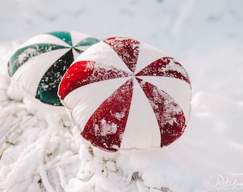 May include: Two round throw pillows with a pinwheel design. The pillow on the left is teal and white, and the pillow on the right is red and white. Both pillows are covered in a light dusting of snow.