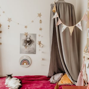 May include: A gray canopy bed with a red velvet bedspread and a cat lying on the bed. The canopy is decorated with a floral garland. There is a rainbow wall hanging and a poster on the wall behind the bed.