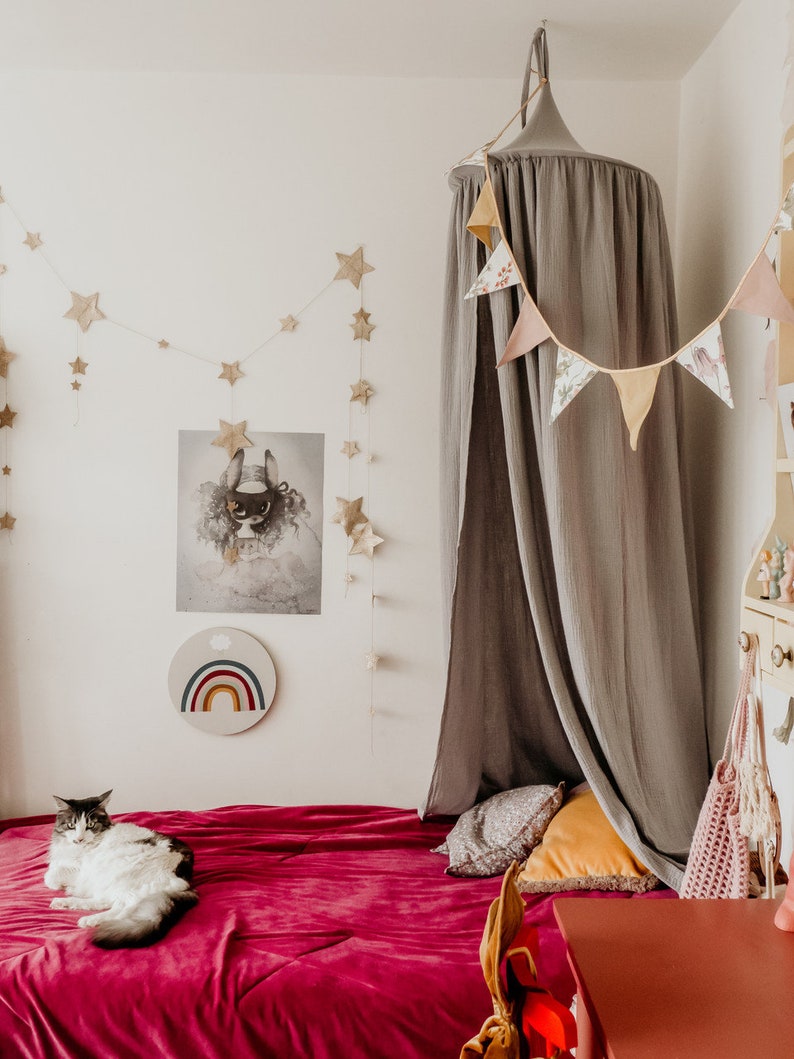 May include: A gray canopy bed with a red velvet bedspread and a cat lying on the bed. The canopy is decorated with a floral garland. There is a rainbow wall hanging and a poster on the wall behind the bed.