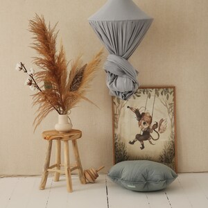 May include: A light gray fabric canopy hanging from the ceiling, with a wooden stool and a vase of dried pampas grass in front of it. A framed illustration of a winged creature on a swing is on the wall behind the stool.