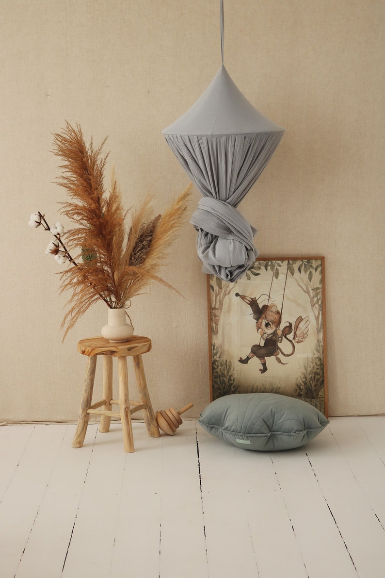 May include: A light gray fabric canopy hanging from the ceiling, with a wooden stool and a vase of dried pampas grass in front of it. A framed illustration of a winged creature on a swing is on the wall behind the stool.