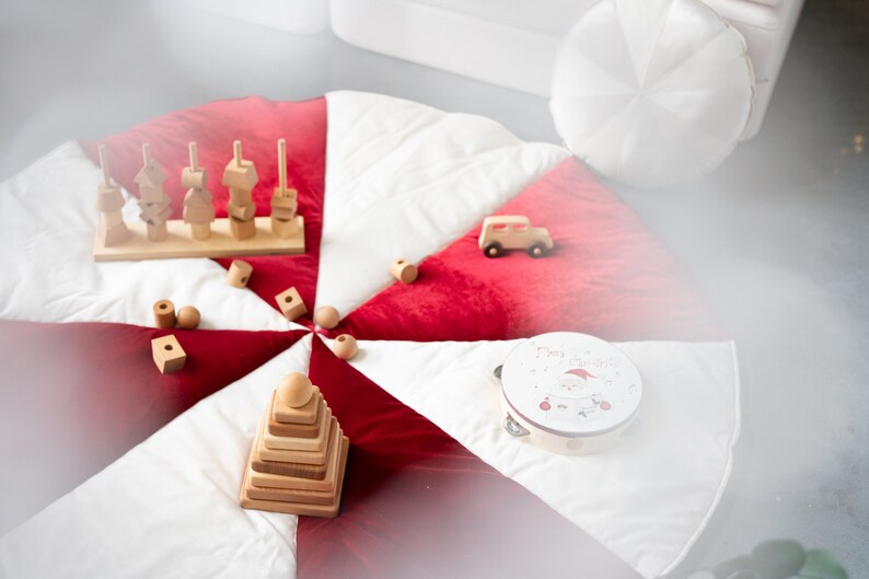 May include: A white and red play mat with a wooden toy stacking pyramid, a wooden toy with different shapes, a wooden toy car, and a tambourine with a Santa Claus design and the text "Merry Christmas".