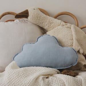 May include: A white fluffy goose plush toy and a light blue cloud-shaped pillow on a white waffle weave blanket. The goose and pillow are on a wooden bed frame.