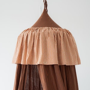 May include: A brown and white gingham canopy with a striped lower section. The canopy has a conical top and a ruffled edge. The fabric is a warm, earthy tone, suitable for a nursery or children's room. The canopy hangs from a white hook.