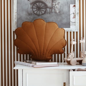 Puede incluir: Una almohada de color marrón acolchada con forma de concha marina se encuentra sobre una cómoda blanca con un fondo de papel tapiz a rayas marrones y blancas. Hay juguetes de madera en la cómoda, incluyendo una jirafa, un ratón y un juguete con forma de estrella.