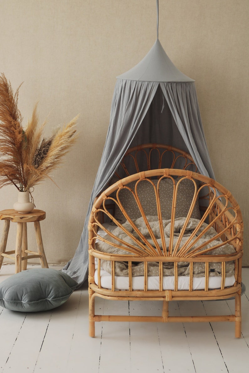 May include: A light gray canopy draped over a natural woven rattan bed frame with a white and gray patterned bedding. A gray velvet pillow sits on the floor in front of the bed.