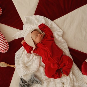 May include: A baby wearing a red onesie and a white bonnet is sleeping on a white and red blanket. The blanket is on a white surface.