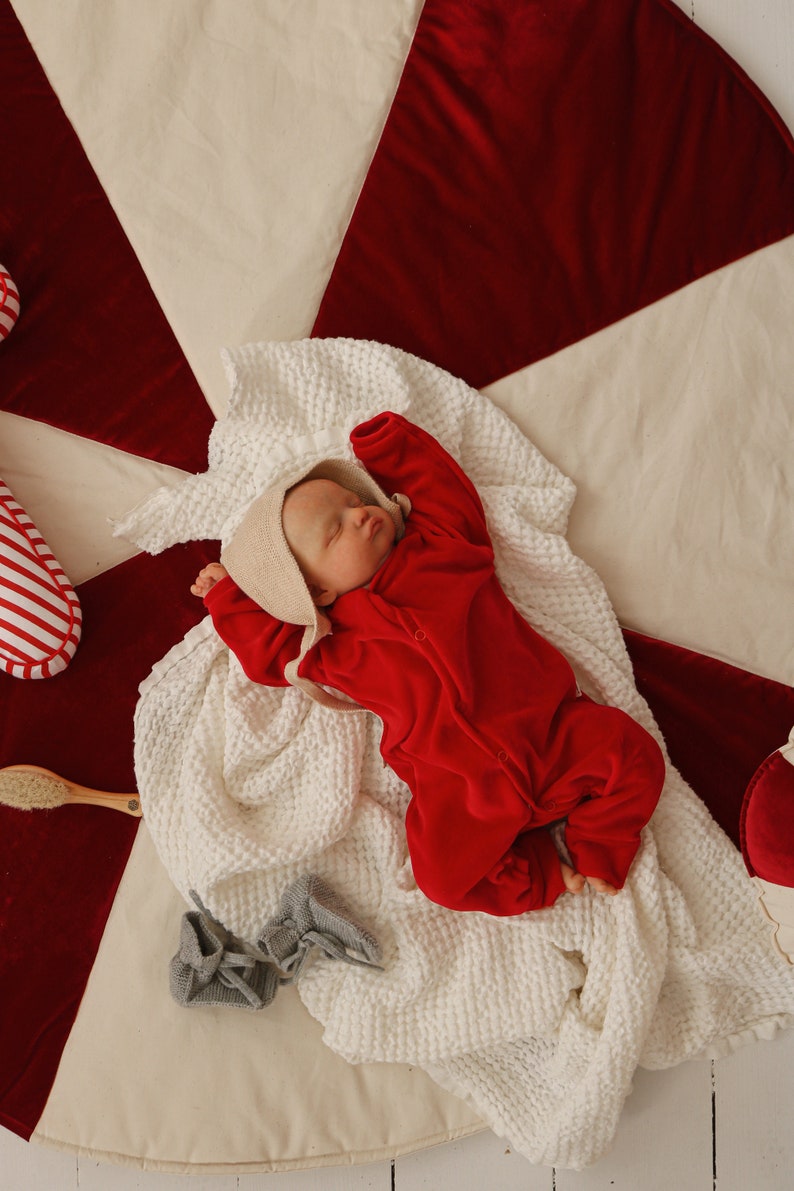 May include: A baby wearing a red onesie and a white bonnet is sleeping on a white and red blanket. The blanket is on a white surface.