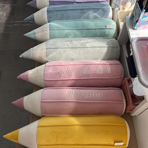 Op de afbeelding: Een verzameling van acht potloodvormige kussens gerangschikt in een rij. Elk kussen heeft een punt van lichtcrèmekleurige stof en een cilindervormig lichaam in diverse kleuren waaronder paars, blauw, groen, roze en geel met geborduurde namen.