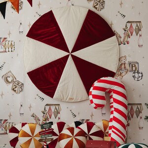 May include: A red and white play mat with a candy cane pillow, a red mailbox with the text "Santa's Mail", and several colourful pillows with a candy cane design. The background features a white wallpaper with a whimsical circus theme.