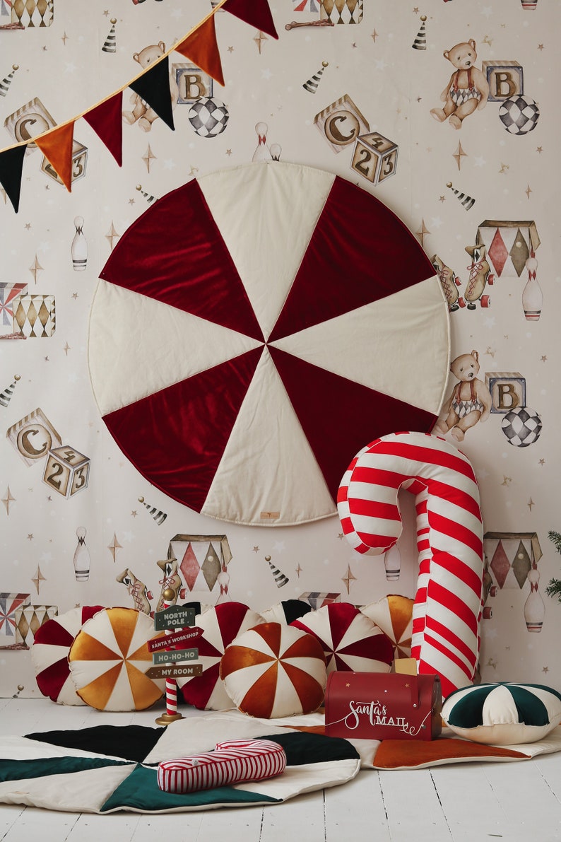 May include: A red and white play mat with a candy cane pillow, a red mailbox with the text "Santa's Mail", and several colourful pillows with a candy cane design. The background features a white wallpaper with a whimsical circus theme.
