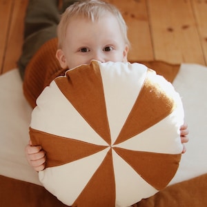 Può includere: Un bambino si nasconde dietro un grande cuscino rotondo marrone e bianco con un design a raggiera. Il cuscino è fatto di tessuto e ha un centro bianco con raggi marroni che si irradiano verso l'esterno.