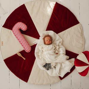 May include: A baby wearing a white knitted outfit and a brown animal hat lies on a cream and red candy cane patterned play mat. A red and white candy cane pillow and a wooden hairbrush are on the mat.