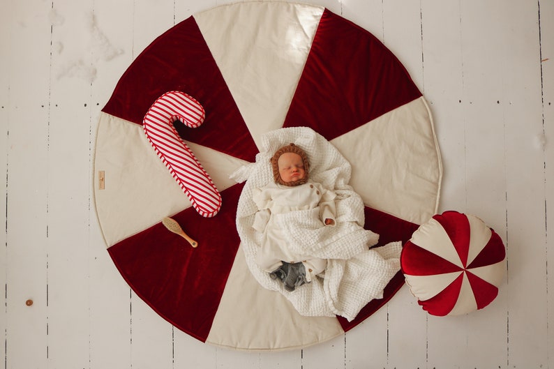 May include: A baby wearing a white knitted outfit and a brown animal hat lies on a cream and red candy cane patterned play mat. A red and white candy cane pillow and a wooden hairbrush are on the mat.